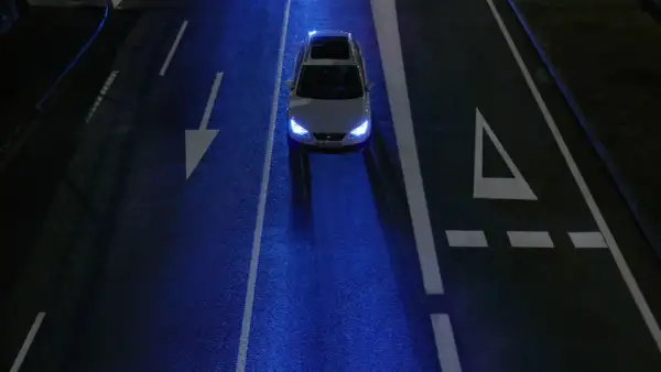 Un coche blanco con las luces encendidas, conduciendo de noche por una carretera oscura con marcas viales blancas y un reflejo azulado.
