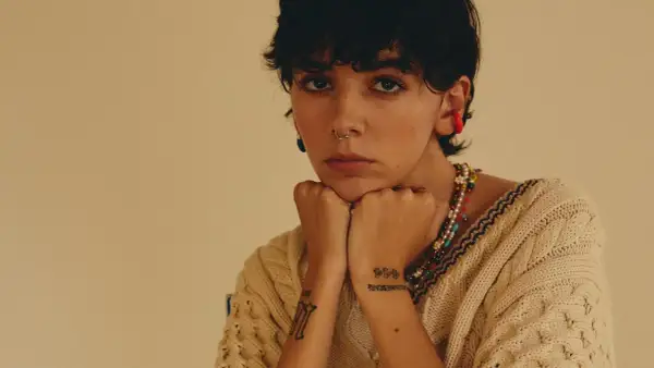 Un'ariete cantante con piercing e tatuaggi, indossa un maglione beige e una collana colorata.