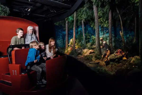 Familj i en vagn på ett vikingamuseum i Sverige, tittar på en dramatisk scen med vikingar i en skog.
