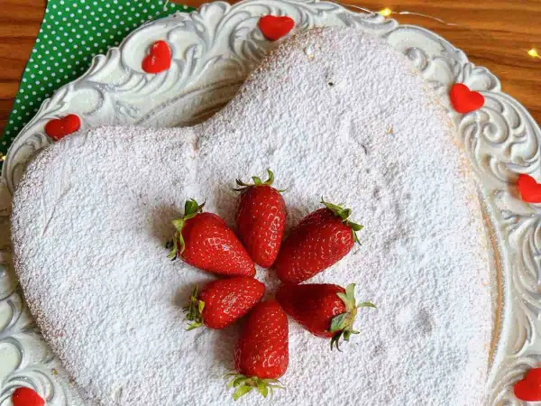 Torta panna e fragole velocissima a forma di cuore, cosparsa di zucchero a velo e decorata con fragole fresche e cuoricini rossi.