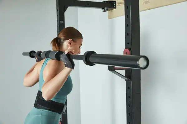 Kobieta w rękawiczkach i pasie treningowym przygotowuje się do przysiadów ze sztangą. Trening push pull legs to jej sposób na siłę.