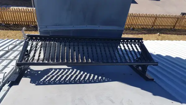 En takbrygga framför en stor ventilationsanläggning, liknande en skorsten. Solen kastar ett mönstrat skuggspel.