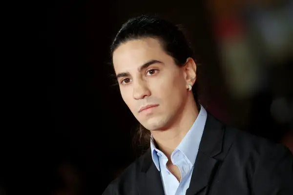 Ritratto di Tedua, rapper italiano, con capelli lunghi legati e un orecchino scintillante. La sua età è difficile da definire, ma emana un'aura di maturità artistica.