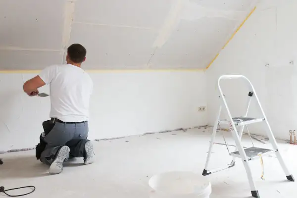 Mann kniet und spachtelt Deckenfugen, um Rigipsplatten zu kleben. Eine Leiter und ein Eimer stehen bereit.