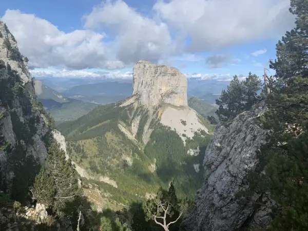 Majestätischer Mont Aiguille ragt aus grünen Tälern empor, umgeben von Felsen und Kiefern unter einem dramatischen Wolkenhimmel.