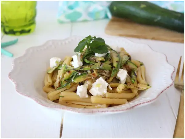 Pasta fredda con zucchine grigliate e cubetti di formaggio fresco, guarnita con erbe aromatiche. Un piatto estivo leggero e gustoso.