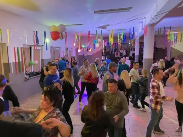 Wesoła zabawa w remizie, ludzie tańczą przy muzyce. Kolorowe serpentyny i balony tworzą świąteczną atmosferę.