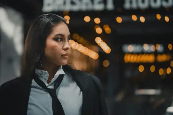 Eine Frau im Business-Casual-Look mit Hemd, Krawatte und Sakko. Der Hintergrund ist unscharf mit Lichtern.