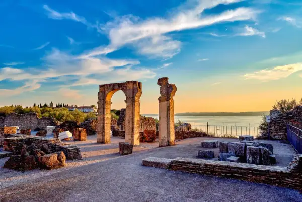Resti archeologici delle Grotte di Catullo con un arco in pietra e colonne, sullo sfondo il lago e un cielo azzurro al tramonto.