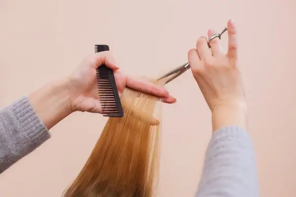 Hände mit Kamm und Schere schneiden blonde Haare. So kannst du kaputte Spitzen selber schneiden und deinem Haar neuen Glanz verleihen.