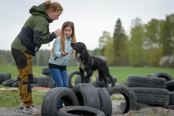 En kvinna och en flicka leker med en svart hundvalp bland gamla däck utomhus.