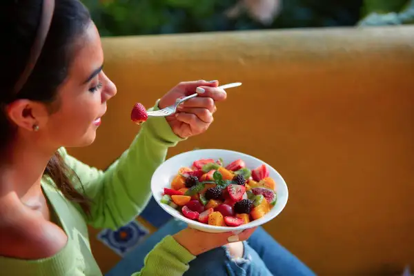 Une jeune femme savoure une salade de fruits colorée, une fraise à la fourchette. La naturopathie suggère quand manger les fruits pour une digestion optimale.