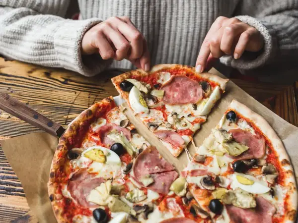 Une personne choisit une part de pizza avec une riche pizza garniture : jambon, œufs, olives, artichauts et champignons.