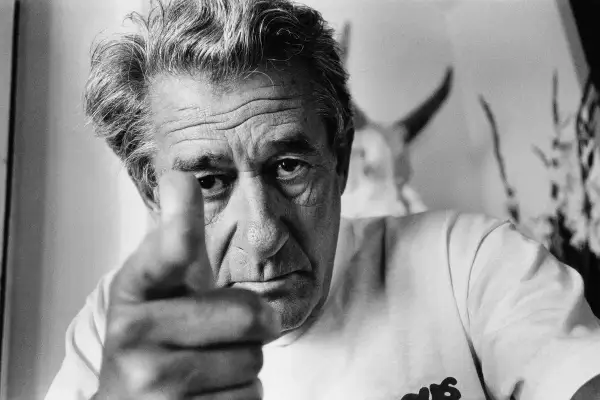 Helmut Newton, z palcem wskazującym w obiektyw, patrzy intensywnie. W tle widać fragmenty jego charakterystycznych prac.