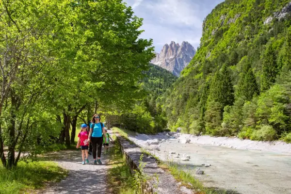 Famiglia in escursione ad Andalo, cammina lungo un sentiero con un fiume cristallino e montagne maestose sullo sfondo.