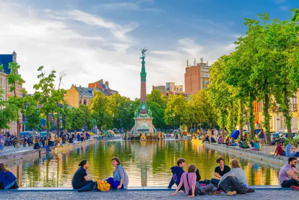 Des gens profitent d'une journée ensoleillée au bord d'un étang, un monument imposant se dresse en arrière-plan. Une belle scène pour visiter Bruxelles.