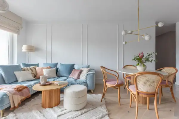 Miękka, błękitna sofa i pudroworóżowe akcenty tworzą przytulną atmosferę. Ciepłe kolory do salonu podkreślają drewniane meble i jasny dywan.