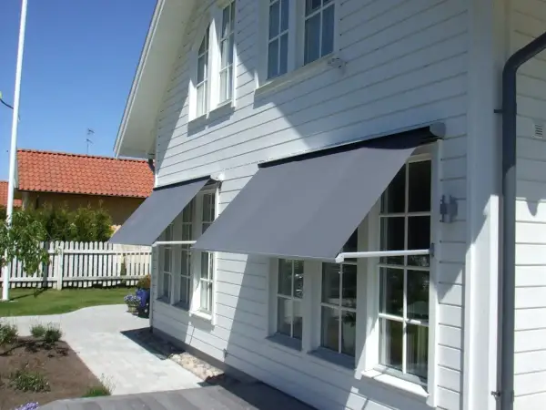 Grå markiser monterade på ett vitt trähus ger skugga åt fönstren.