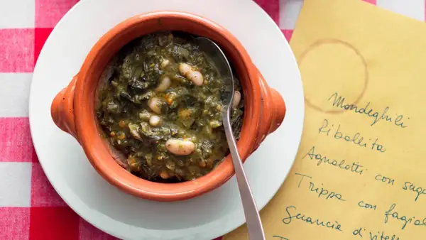 Zuppa di cavolo nero e fagioli in ciotola di terracotta, servita su un piatto bianco con tovaglia a quadri rossi. Accanto, un foglio giallo con un elenco di piatti.