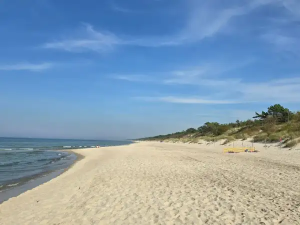 Słoneczny dzień nad zalewem kamieńskim. Szeroka, piaszczysta plaża z łagodnymi falami i zielonymi wydmami w tle. Kilka osób odpoczywa na piasku.