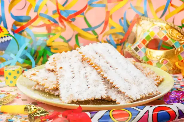 Bugie ricetta originale, dolci fritti cosparsi di zucchero a velo, su un piatto con coriandoli e maschere di Carnevale.