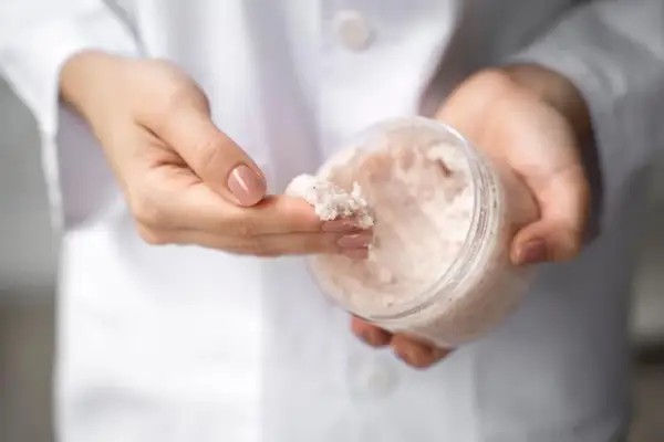 Dłonie w białym kitlu pobierają z pojemnika drobnoziarnisty peeling. To świetny sposób, by dowiedzieć się, co to mocznik i jak działa na skórę.