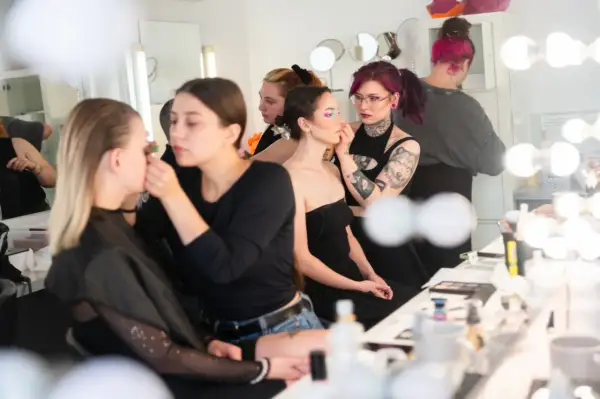 Charakteryzatorka filmowa pracuje nad makijażem modelki. W tle inne osoby przygotowują się do sesji.