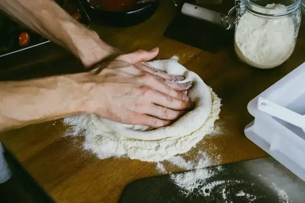 Dłonie formują ciasto na pizzę na drewnianym blacie. W tle słoik z mąką i sos pomidorowy. Zastanawiasz się, ile ma rosnąć ciasto na pizzę?