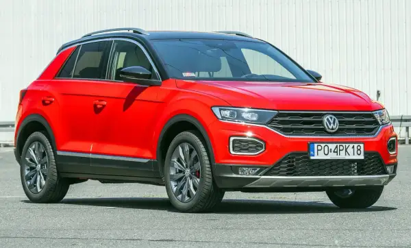 Czerwony, używany Volkswagen T-Roc z czarnym dachem i przyciemnianymi szybami, zaparkowany na tle białej ściany.