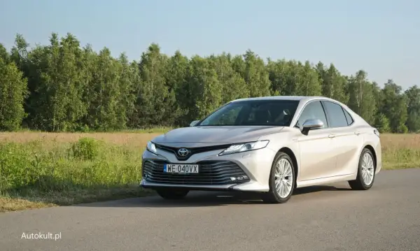 Elegancka Toyota Camry 2025 w kolorze szampańskim na tle zielonego krajobrazu.