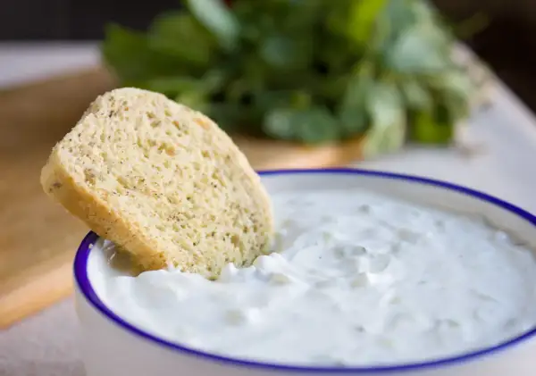Ein Stück Brot wird in eine Schüssel mit cremigem, selbstgemachtem Zaziki getunkt. Frische Kräuter im Hintergrund.