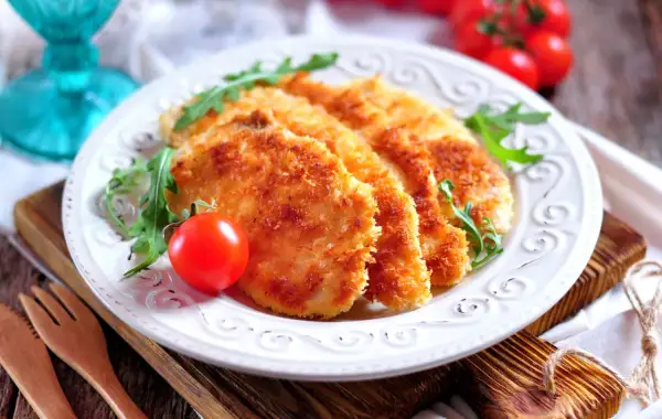 Delizioso pollo impanato al forno, croccante e dorato, servito con pomodorini e rucola su un piatto bianco.