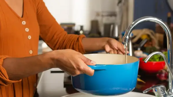 Une personne remplit une cocotte bleue sous un robinet pour faire bouillir de l'eau chaude.
