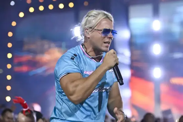 Artysta z blond włosami i niebieskimi okularami śpiewa na scenie. W tle widać światła i tłum. To koncert na **Skolim stacja**.