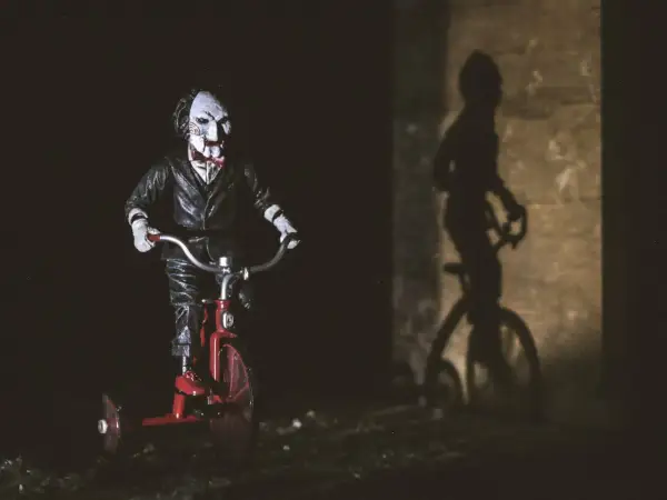 Lalka Billy z Piły na czerwonym trójkołowcu, cień na ścianie. Idealne do listy horrorów.