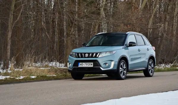 Jasnoniebieska Suzuki Vitara na drodze leśnej, z drzewami w tle i resztkami śniegu.