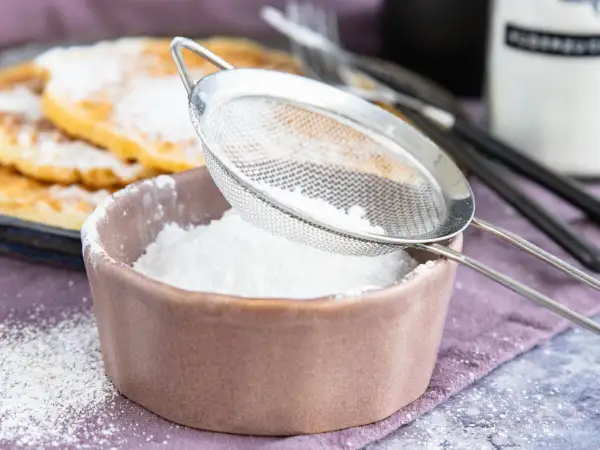 Feiner Puderzucker rieselt aus einem Sieb in eine Schale. So einfach lässt sich puderzucker selber machen für leckere Waffeln.