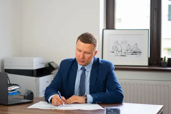 Mężczyzna w garniturze siedzi przy biurku i pisze na dokumentach.