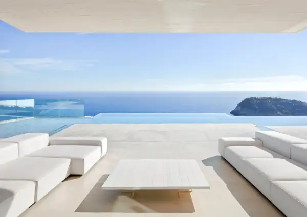 Terrasse moderne avec canapés blancs et table basse, bordée par une piscine à débordement et la mer bleue. Le **béton ciré autour de la piscine** crée une ambiance épurée et luxueuse.