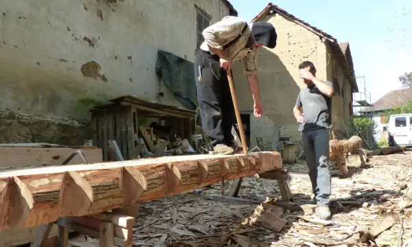 Deux hommes travaillent sur un tronc d'arbre, pratiquant l'équarrissage bois avec une hache. Le bois est façonné avec soin.