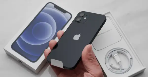 Czarny iPhone 12 w ręce, obok pudełko z telefonem i kablem. Chcesz wiedzieć, jak resetować iPhone?