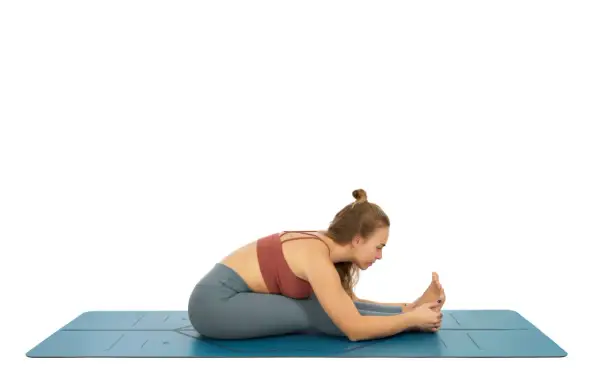 Mujer en esterilla azul, practicando una de las variantes de uttanasana, con las piernas estiradas y el torso inclinado hacia adelante, sujetando los pies.