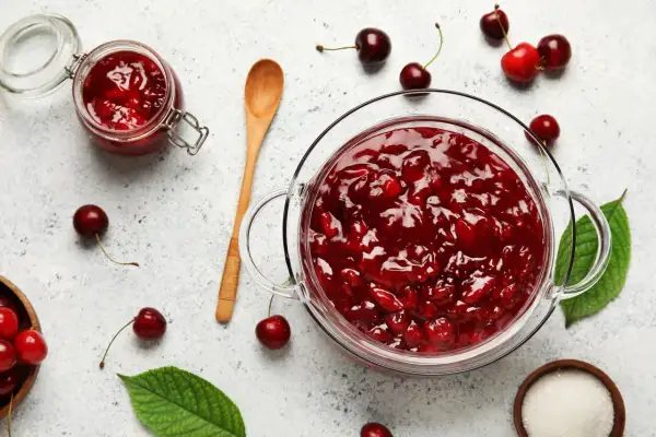Un bocal et un bol en verre débordent de confiture de cerises maison. Des cerises fraîches et des feuilles vertes parsèment la surface.