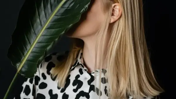 Blond włosy lśniące jak po zabiegu z żelatyną na włosy. Kobieta w bluzce w panterkę ukryta za dużym, zielonym liściem.