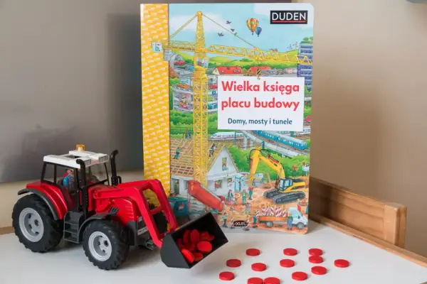 Czerwony traktor z ładowarką i rozsypanymi czerwonymi krążkami obok książki "Wielka księga placu budowy".