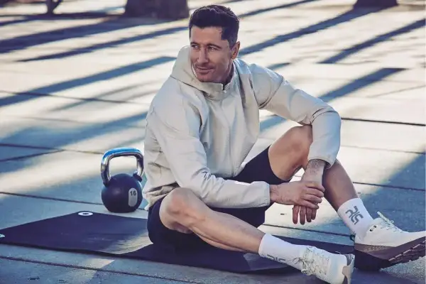 Robert Lewandowski w sportowym stroju, siedzi na macie treningowej obok odważnika. Ciekawe informacje o jego karierze i treningach.