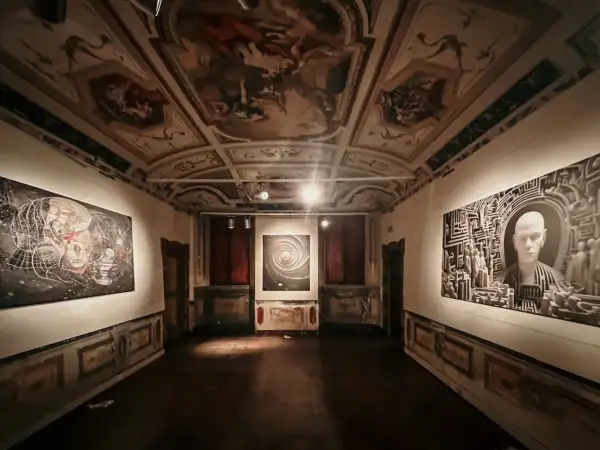 Sala di una galleria d'arte con soffitto affrescato e opere moderne. Le mostre ad Ancona presentano un'installazione con un volto stilizzato e un'opera astratta.