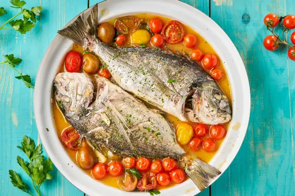 Orata all'acqua pazza, due pesci interi cotti in un brodo saporito con pomodorini e olive, guarniti con prezzemolo fresco.