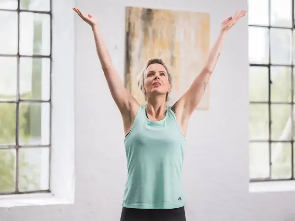 Femme aux bras levés, profitant d'un moment pour calmer son mental. La lumière naturelle baigne la pièce, accentuant sa sérénité.