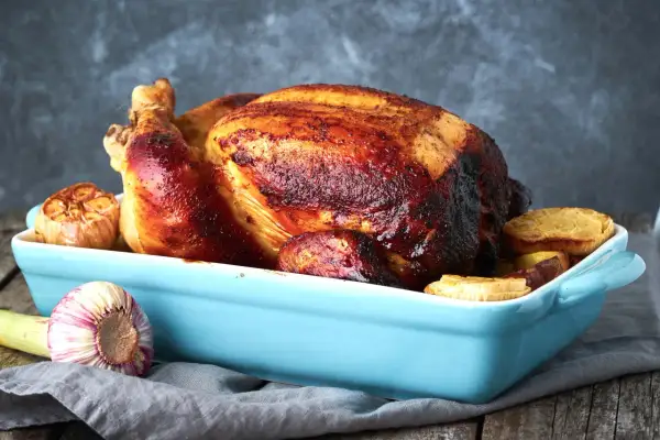 Un succulento pollo al forno dorato e croccante, servito in una teglia azzurra con aglio arrosto e patate.